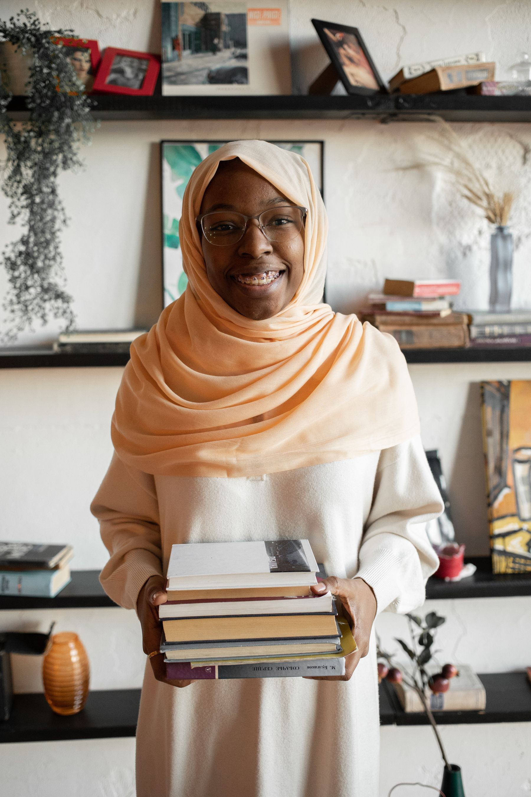 A smiling woman holding a stack of books in front of bookshelves